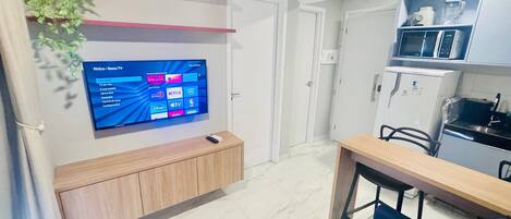 A modern living room with a flat-screen TV mounted on the wall, a wooden cabinet, a dining table with chairs, and a kitchen area with a microwave and refrigerator.