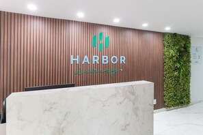 Balcão de recepção com painel de parede de madeira, logotipo com 'HARBOR' e 'Luzurg', e parede verde com plantas.