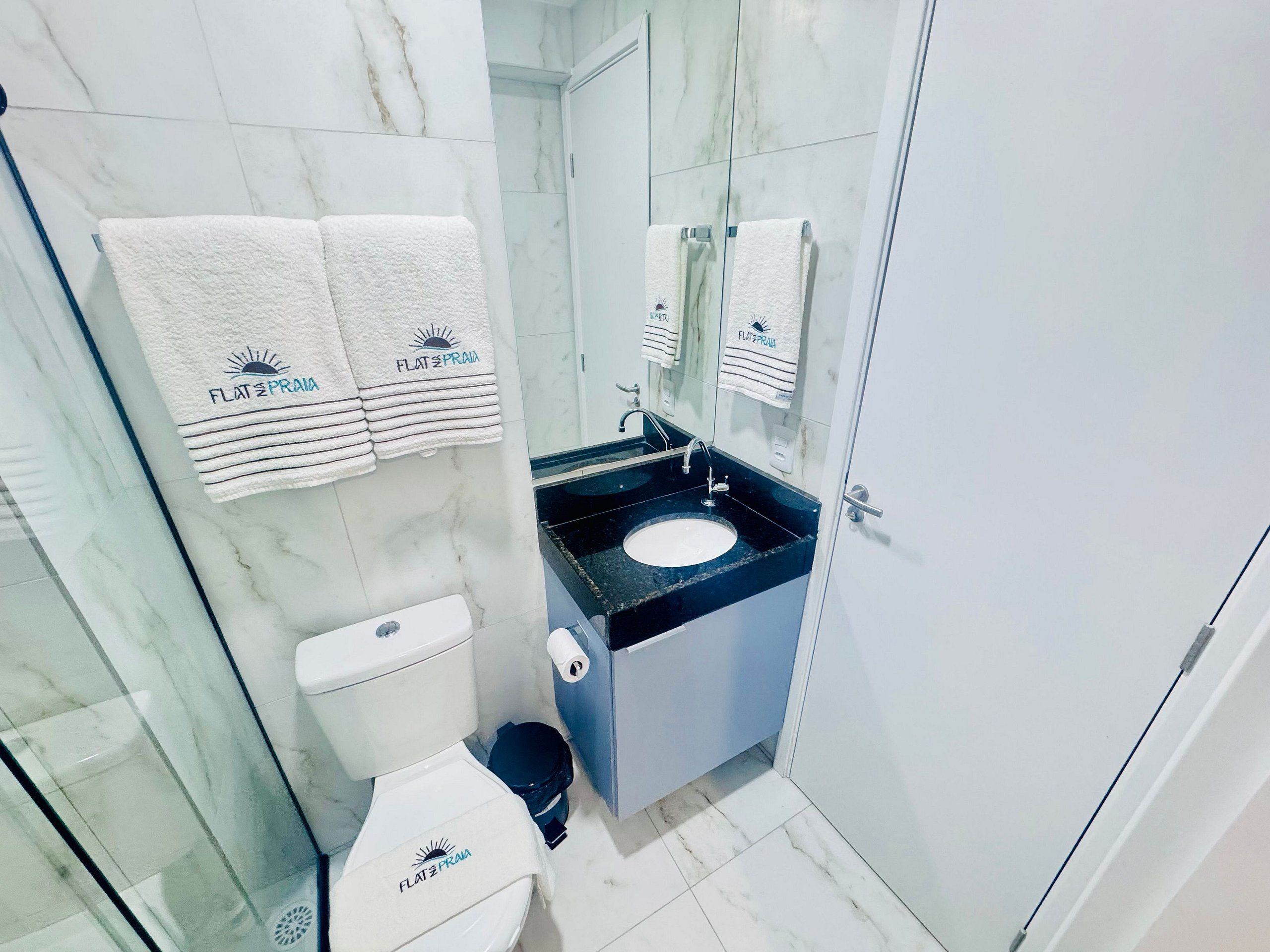 Une salle de bain avec des toilettes, un lavabo et un miroir. On y trouve des serviettes portant la marque « FLAT & PRAIA ».
