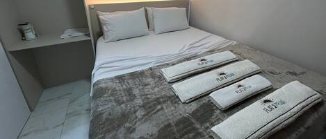 A hotel room with a bed, pillows, a bedside table with a phone, an air conditioning unit, and towels with a logo.