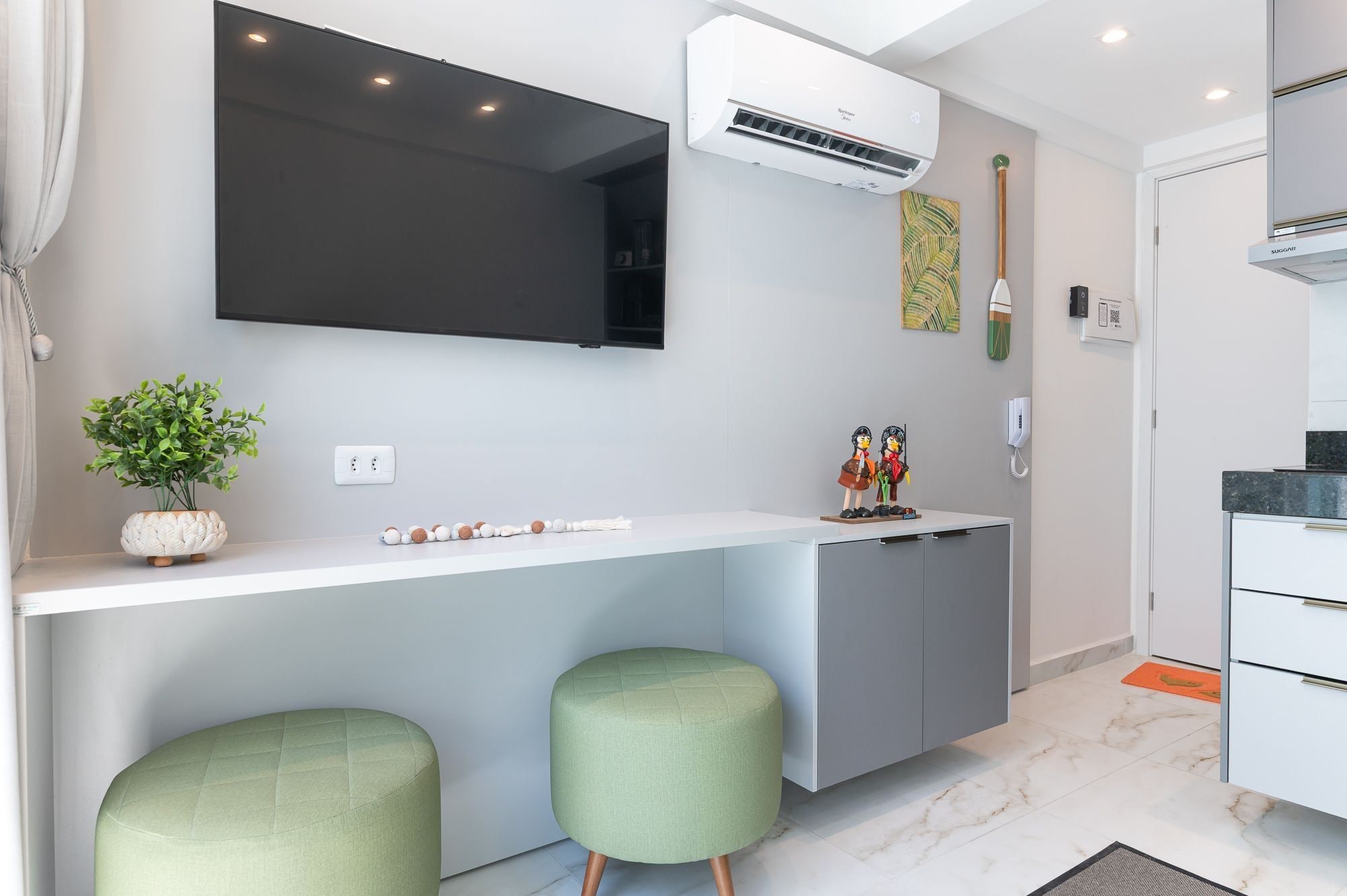 Una cocina moderna con encimera blanca, dos taburetes verdes, un equipo de aire acondicionado y un televisor montado.