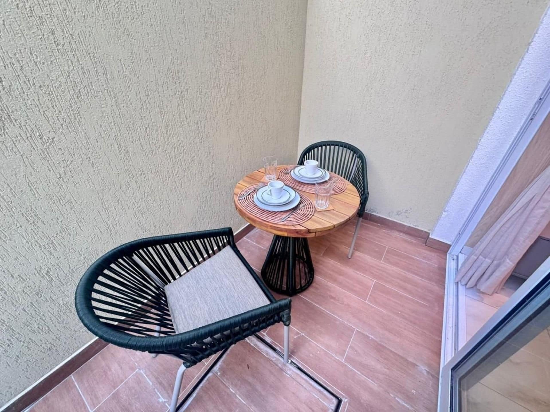 Een klein balkon met een ronde tafel gedekt voor twee personen, twee zwarte stoelen en een glazen deur naar binnen.