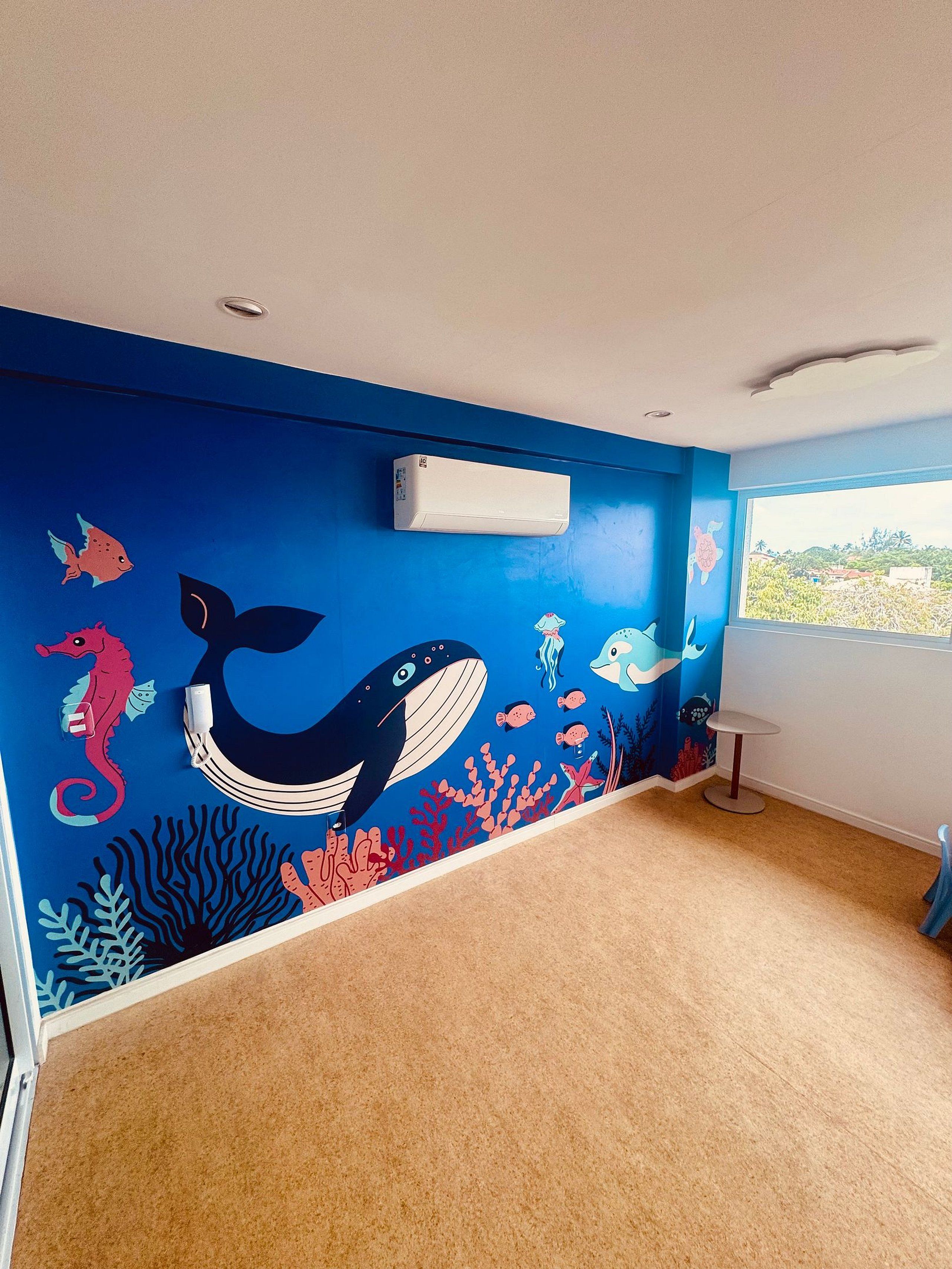 Une pièce avec une fresque murale bleue représentant la vie marine, un climatiseur blanc et une fenêtre offrant une vue.
