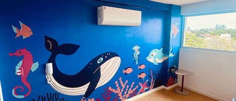 Une pièce avec une fresque murale bleue représentant la vie marine, un climatiseur blanc et une fenêtre offrant une vue.