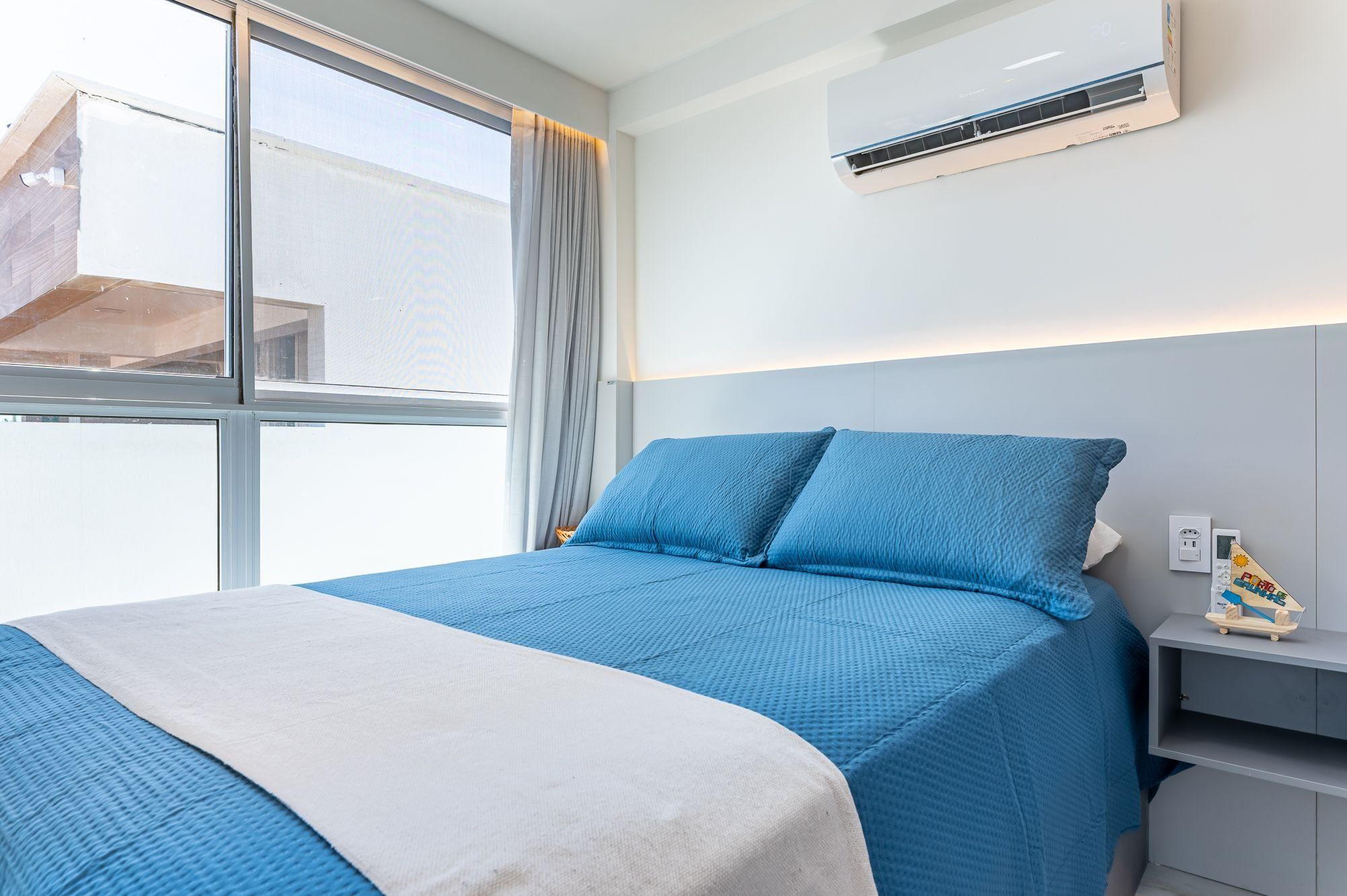 A bedroom with a bed, a window, an air conditioning unit, and a small shelf with a toy.