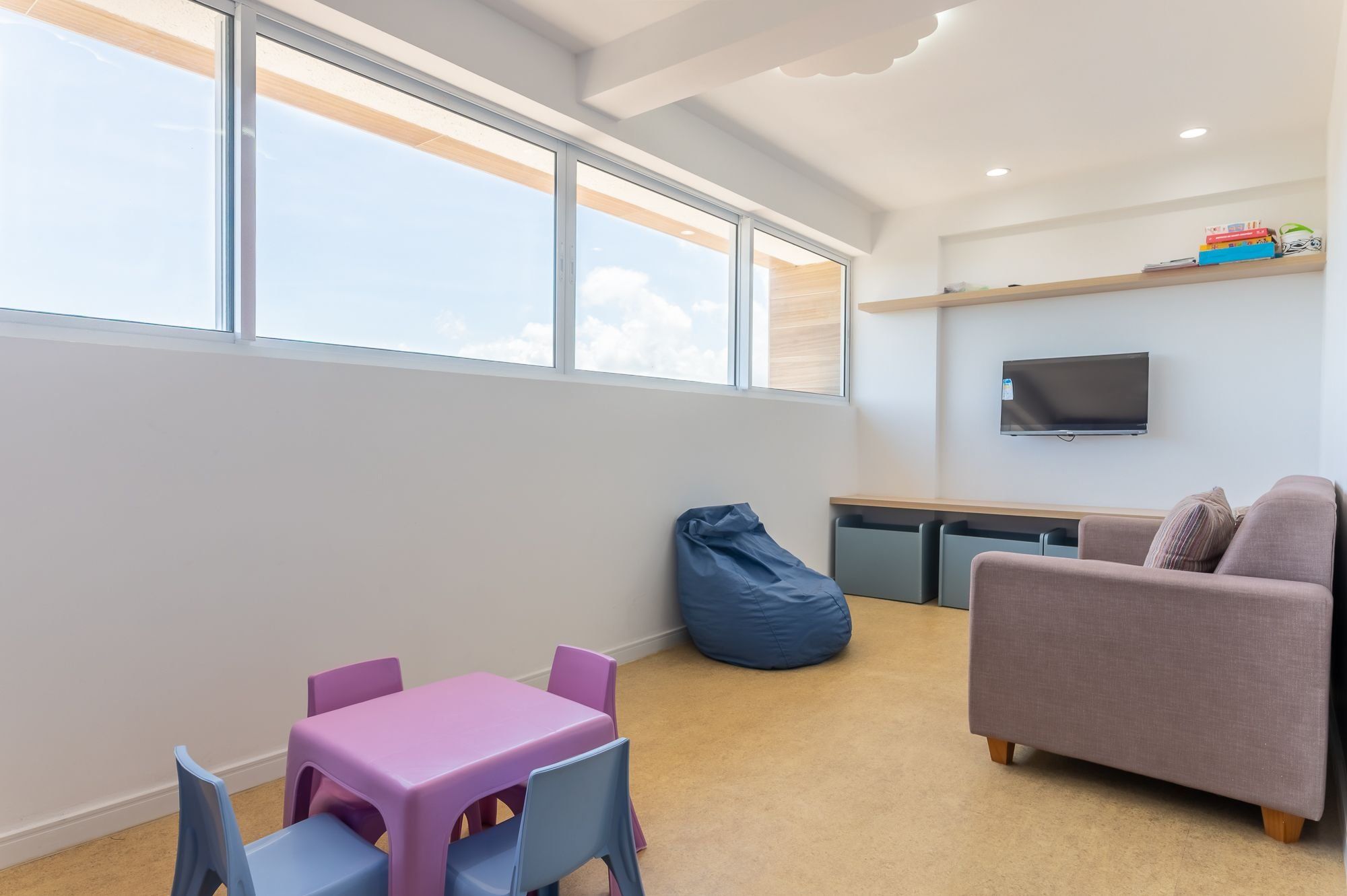 Una habitación moderna con un ventanal grande, una mesa rosa con sillas, un sillón orejero azul, un sofá gris y un televisor de pantalla plana en la pared.
