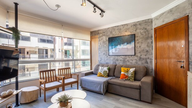 A modern living room with a grey sofa, wooden chairs, a coffee table, and a large window offering a view of the city. - Apt With Balcony and BBQ 30m From the sea Rdm0101 (Itapema)