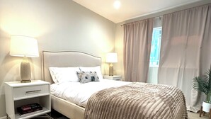 A modern bedroom with a large bed, two bedside tables with lamps, a nightstand, a potted plant, and a window with curtains. - Classy 4-bedroom house with AC in Redondo Beach (Redondo Beach)
