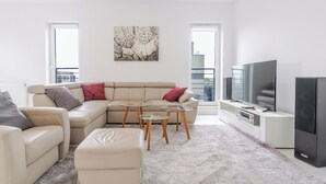 A modern living room with a beige sectional sofa, a glass coffee table, a small ottoman, a flat-screen TV, and a large window with a view of a building. - Francuska Comfort Apartment (Katowice)
