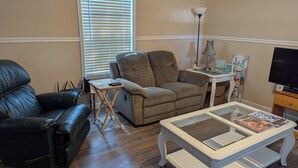 A living room with a black leather sofa, a grey armchair, a white coffee table, and a television on a wooden stand. - Lovely 4-bedroom mobile home in Oak Island 6 MIN WALK TO BEACH (Oak Island)