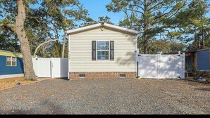 A single-story mobile home with a white exterior, a front garage door, and a gravel driveway. - Lovely 4-bedroom mobile home in Oak Island 6 MIN WALK TO BEACH (Oak Island)