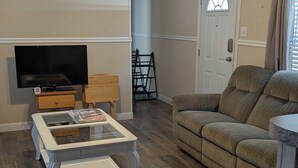 A living room with a ceiling fan, a flat-screen TV, a glass coffee table, a sofa, and a door with a window. - Lovely 4-bedroom mobile home in Oak Island 6 MIN WALK TO BEACH (Oak Island)