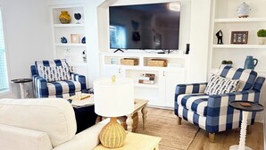 Un salon avec un canapé blanc, des fauteuils à carreaux bleus et blancs, une table basse en bois et un téléviseur à écran plat fixé à un mur blanc.