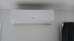 A white GREE air conditioning unit mounted on a wall in a room with a plain white wall and a black curtain.
