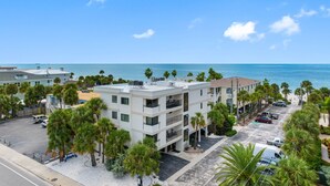 A multi-story hotel with a parking lot, surrounded by palm trees and a view of the ocean.