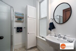 Ein Badezimmer mit weißer Tür, einem Fenster mit Jalousien, einem Waschbecken mit Spiegel und einem Handtuchhalter.