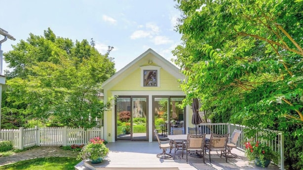 A yellow house with a deck, surrounded by lush greenery and a white picket fence.