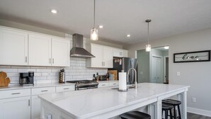 A modern kitchen with white cabinets, a large island with bar stools, a coffee maker, and a stainless steel range hood. - Deer Haven in Georgetown Township (Jenison)