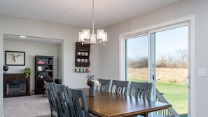 A dining area with a wooden table, six chairs, a chandelier, a fireplace, a bookshelf, and a large window with a view of the outdoors.