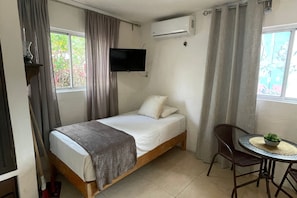 Uma cama de solteiro com uma televisão montada acima, uma pequena mesa redonda com uma planta e uma janela com cortinas.