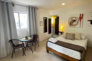 A hotel room with a bed, a small table with chairs, a window with curtains, a wall-mounted artwork, and a nightstand with a lamp. - 05 Conchi Abajo in Isla Mujeres (Isla Mujeres)
