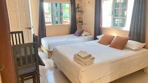 Quarto de hotel com duas camas, uma escrivaninha e uma cadeira. As janelas têm cortinas e há uma prateleira com livros e uma luminária.