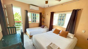 Quarto com duas camas, uma escrivaninha e uma cadeira. Há uma janela com cortinas, um ar condicionado e uma pequena prateleira com objetos.