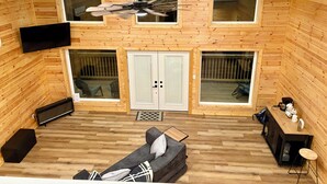 Amplia habitación tipo loft con techo alto, paredes de madera, ventilador de techo, ventana grande, sofá, televisor y pequeña cocina.