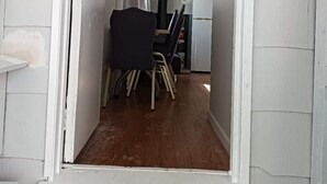 Een smalle gang met witte wanden en een houten vloer die leidt naar een kamer met een donkere stoel en een witte koelkast.