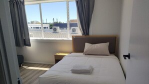 A neatly made bed with white linens and a folded towel, a bedside table with a lamp, and a window with curtains.