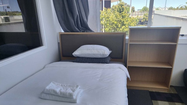 A neatly made bed with a pillow and a folded towel, a wooden shelf, and a window with curtains. - 6 Beds including 1 DBL Bed, 1 Single Bed, 4 Singgle Sofa Bed in 1 Room & 1 Lounge (Auckland)