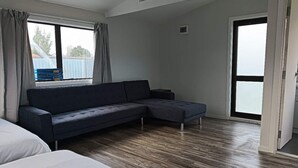 A modern room with a dark grey sectional sofa, a large window with curtains, and a wooden floor. - 4 double and 2 Sofa Beds chalet (Rotorua)