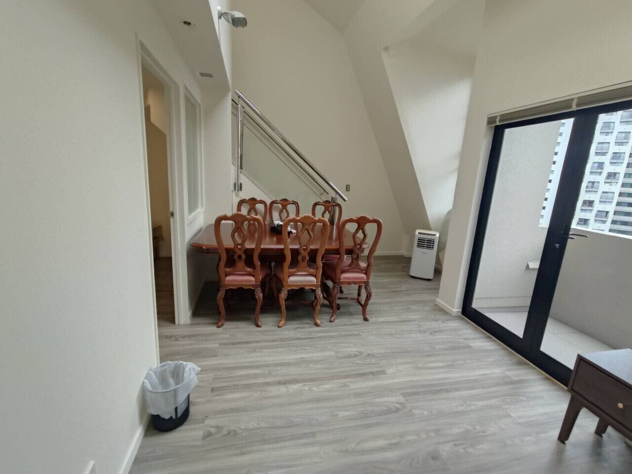 A dining area with a wooden table and chairs, a trash bin, and a sliding glass door leading to a balcony.