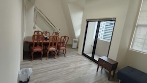 Een kamer met hoge plafonds, een eethoek met een houten tafel en stoelen, een klein bankje en een glazen schuifdeur naar een balkon.