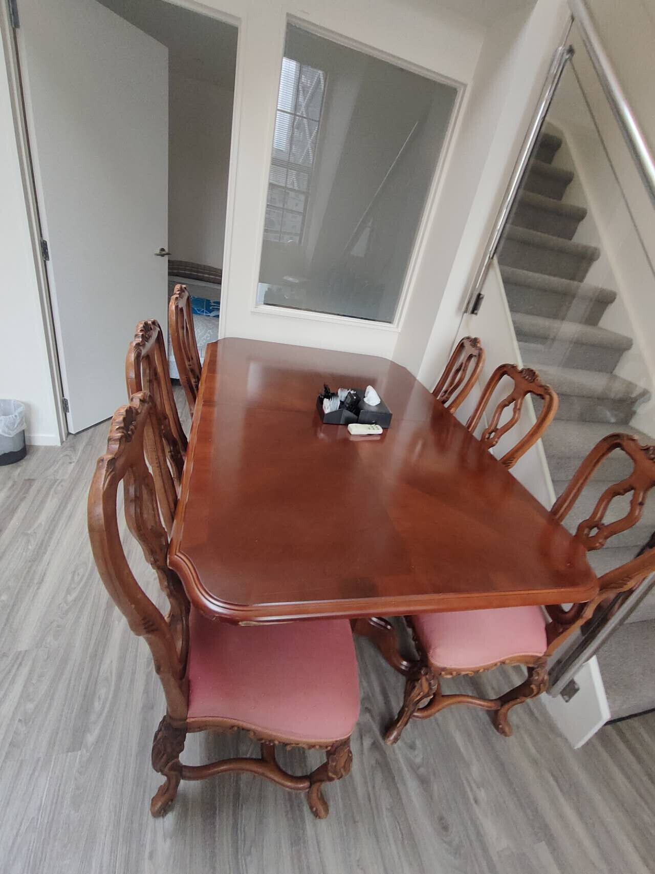 A dining room with a wooden table and chairs, a glass door leading to a staircase, and a trash bin.