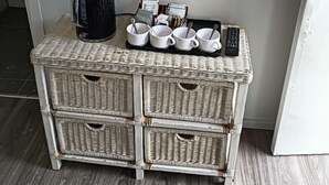 Una mesita auxiliar de mimbre con un hervidor eléctrico negro, tazas blancas, cucharas y un control remoto.