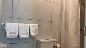 Une salle de bain avec des toilettes, une poubelle et un rideau de douche. On y trouve trois serviettes sur lesquelles est inscrit le mot « AWA ».