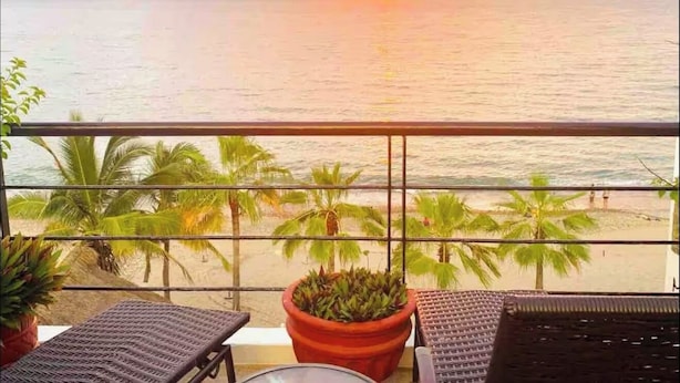Un balcon offrant une vue sur le coucher de soleil sur l’océan, avec des palmiers et une plante en pot.