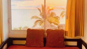 Une chambre dotée d’une grande fenêtre donnant sur une plage au coucher du soleil, avec deux oreillers sur le lit.