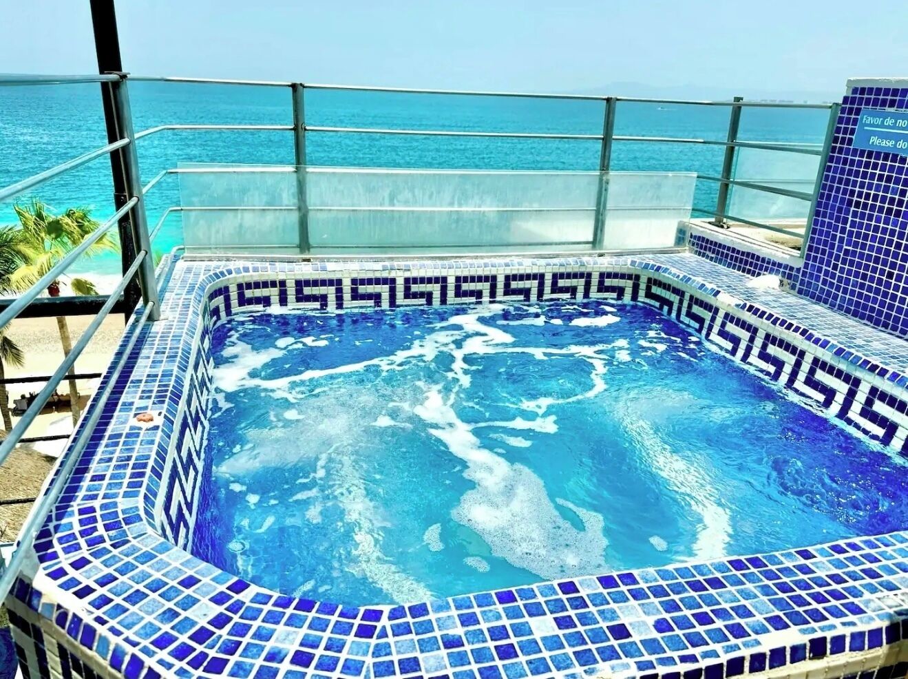 A hot tub with a blue mosaic design, surrounded by a glass railing, overlooking a beach.