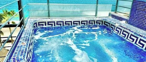 A hot tub with a blue mosaic design, surrounded by a glass railing, overlooking a beach.