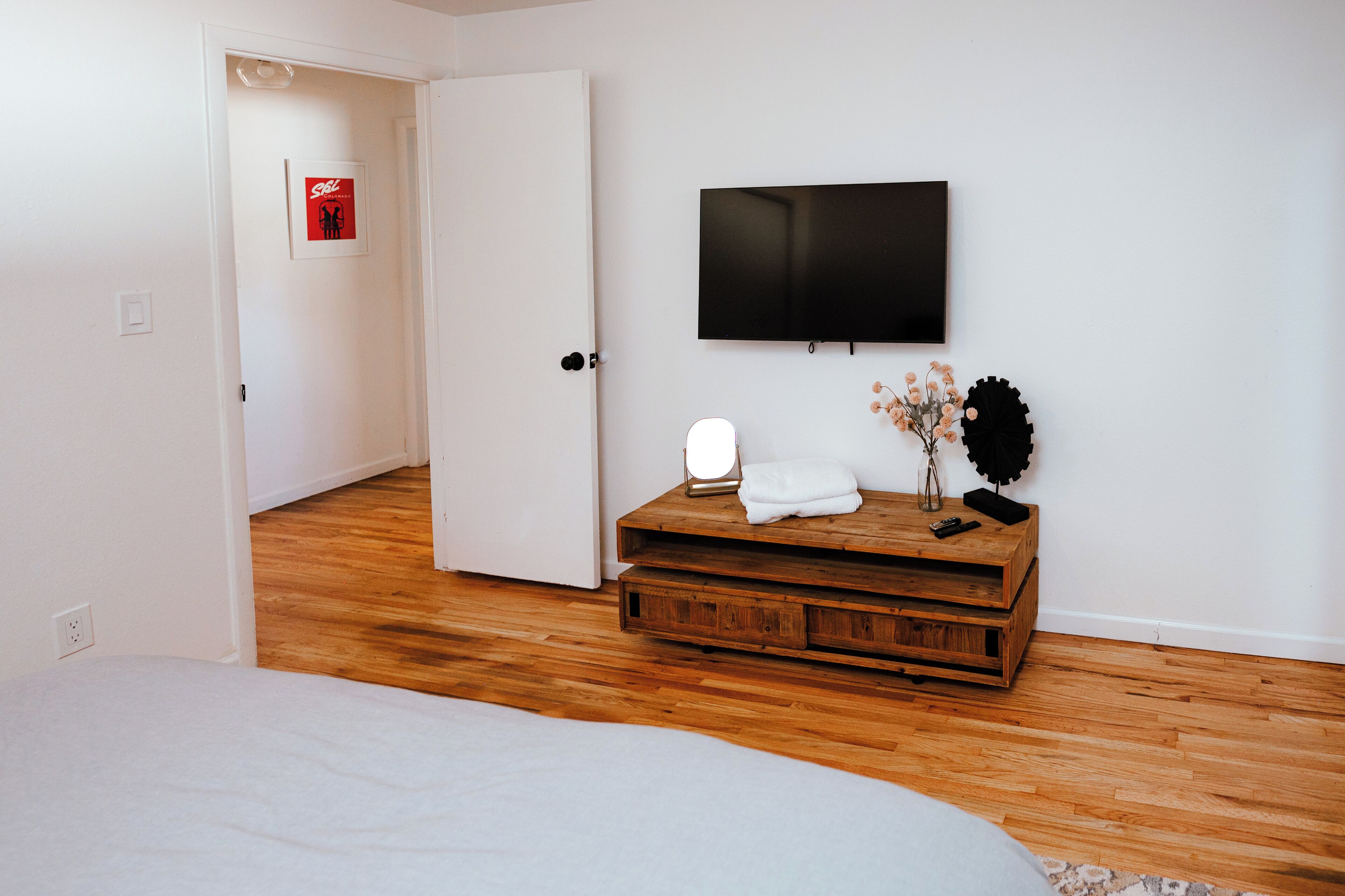A bedroom with a bed, a wooden nightstand with a TV, a wall-mounted light, and a door leading to another room.