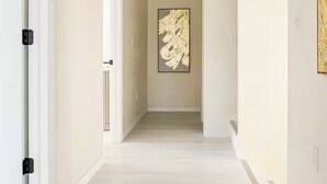 A hallway with a light-colored floor, white walls, and a framed abstract painting on the wall.