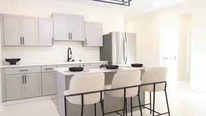 A modern kitchen with grey cabinets, a stainless steel refrigerator, and a bar area with white chairs.