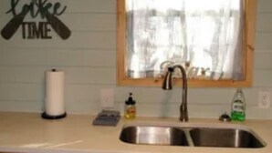 Una cocina con armarios de madera, un lavamanos doble, una ventana con cortinas y un cartel en la pared que dice "ON TIME TIME".