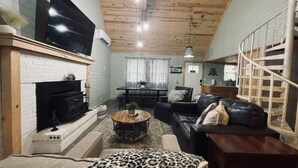 Una sala de estar acogedora con chimenea, un televisor montado en la pared, una mesa de comedor con sillas y una escalera.