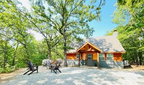 A cabin with a gravel driveway, a covered grill, and two Adirondack chairs.