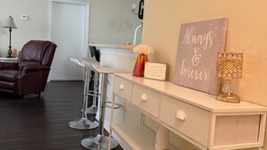 A modern living room with a white console table, a brown leather chair, a white sofa, and a kitchen area with a sink and cabinets.