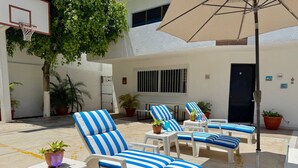 A sunlit outdoor seating area with striped lounge chairs and a large umbrella, surrounded by potted plants and a basketball hoop.