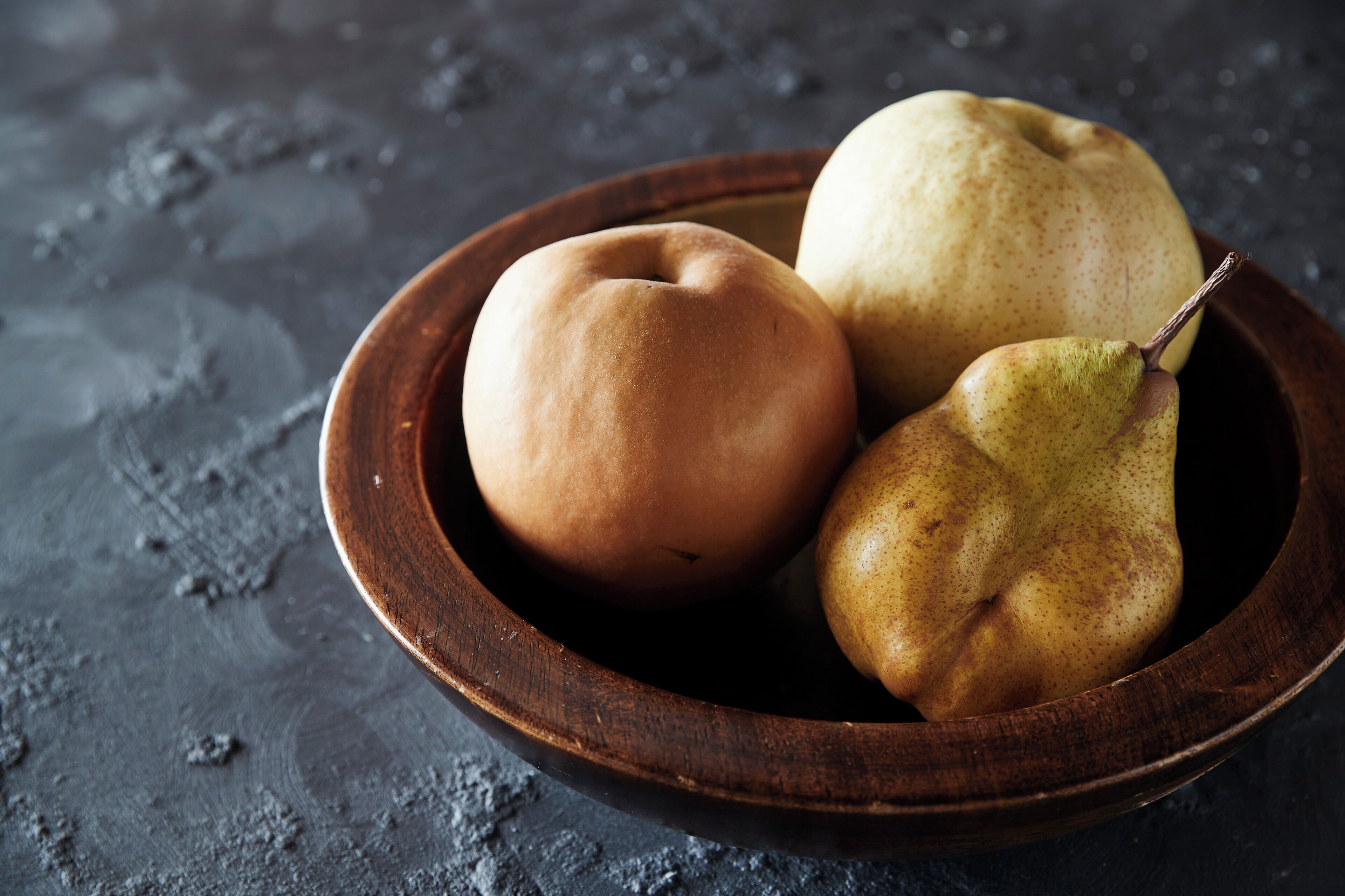 A wooden bowl containing three fruits on a textured surface.